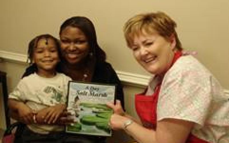 Three smiling people, an adult and a child holding a book, and a woman sitting nearby.