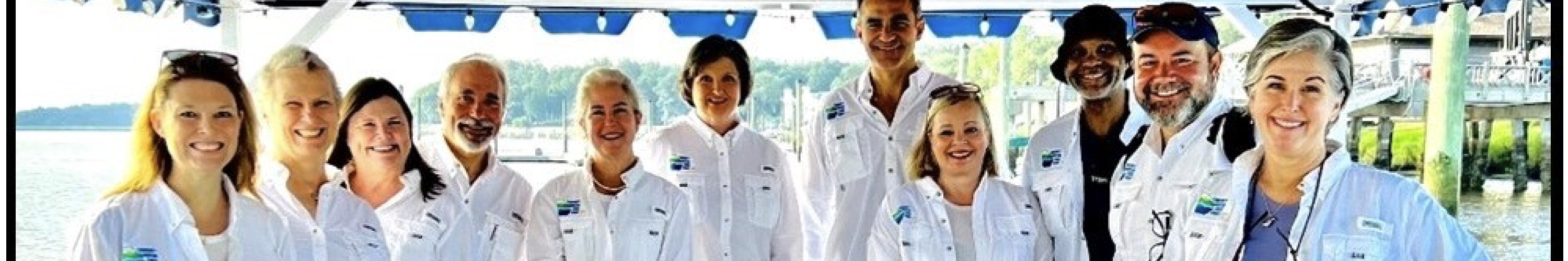 Group of people on a boat, wearing white shirts, smiling under a blue-striped canopy.